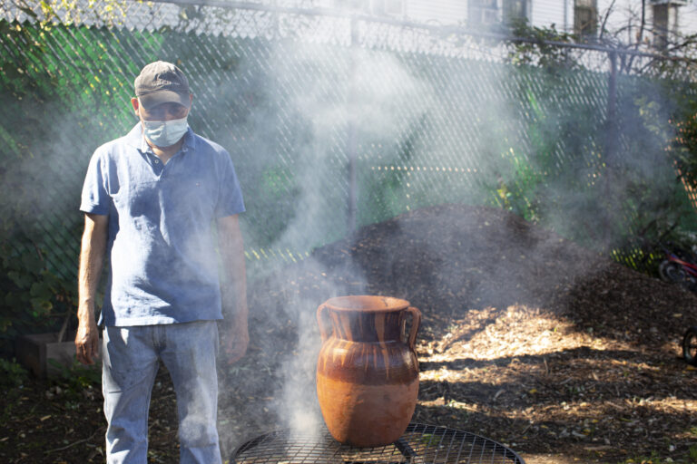 Antonio, who is wearing a light blue shirt, jeans, a baseball cap, and medical mask, stands near a fire. There’s a big pot made out of clay on top of the fire; smoke comes out of the fire. The mounds of soil in the background reflect the morning sunlight.