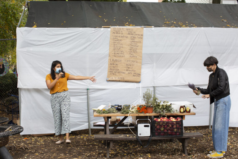 Carolina, wearing an orange shirt, and patterned pants, and Angeles, wearing a black leather jacket and blue jeans, stand in front of the participants of the workshop (out of frame). Angeles has a branch of lavender in her hands; Carolina is speaking using a microphone. Both wear face masks. In the back, a paper kraft board shows a list of words in English and Spanish that Carolina uses as part of the activity (for instance––tierra/earth; memoria/memory, recalling; lengua materna/mother tongue). The table showcases the different plants participants will learn about during the workshop, and jars of honey. There is also a crate with apples, a box with pan de muerto participants will share at the end, and a speaker. Photo by Cinthya Santos-Briones for Brewing Memories workshop, October 24, 2020.