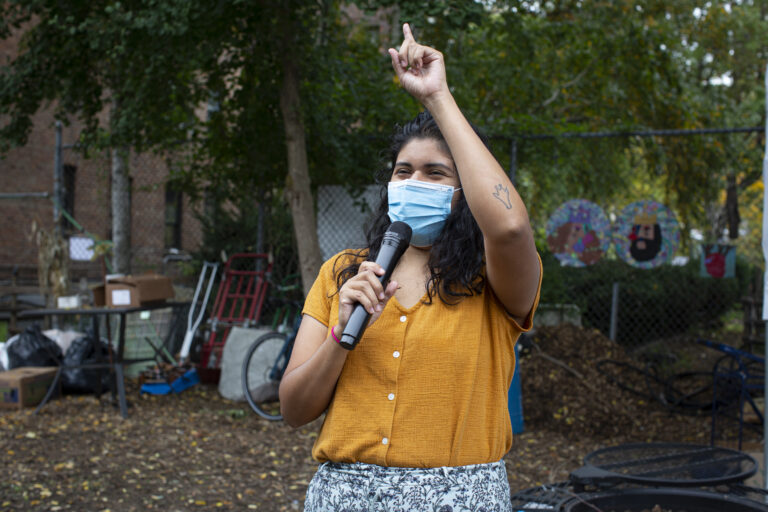 Carolina, wearing an orange shirt and patterned pants, speaks through a microphone she holds with her right hand, while her left arm is up. She wears a medical mask, but one can tell she is smiling. Photo by Cinthya Santos-Briones for Brewing Memories workshop, October 24, 2020.