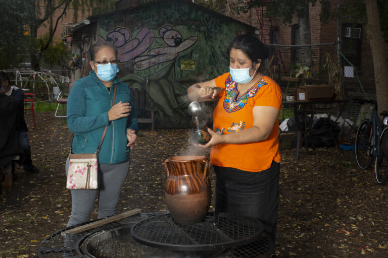 Natalia, wearing an Indigenous Mexican embroidered shirt (orange with flowers), a long black skirt, stands near a fire, serving tea to a workshop participant who is wearing a green jacket and grey pants. Both wear medical masks. Photo by Cinthya Santos-Briones for Brewing Memories workshop, October 24, 2020.