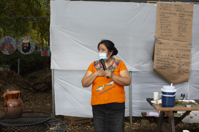 Natalia, wearing an Indigenous Mexican embroidered shirt (orange with flowers), a long black skirt, and a medical mask, shares a memory with the participants of the workshop (out of frame). She uses a microphone to speak, and stands between the table and the fire. In the back, a paper kraft board shows a list of words in English and Spanish that Carolina uses as part of the activity (for instance––tierra/earth; memoria/memory, recalling; lengua materna/mother tongue). Photo by Cinthya Santos-Briones for Brewing Memories workshop, October 24, 2020.