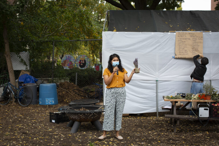 Carolina, wearing an orange shirt, patterned pants, and a medical mask, teaches the properties of lavender. She holds the branch of lavender on her left hand. She wears a medical mask. She stands between the fire pit and the table showcasing the different plants participants will learn about during the workshop. On the bench there is a crate with apples and a speaker. In the back, Angeles, wearing a black jacket and jeans, tries to fix the paper kraft board that shows a list of words in English and Spanish that Carolina will use as part of the activity. Photo by Cinthya Santos-Briones for Brewing Memories workshop, October 24, 2020.