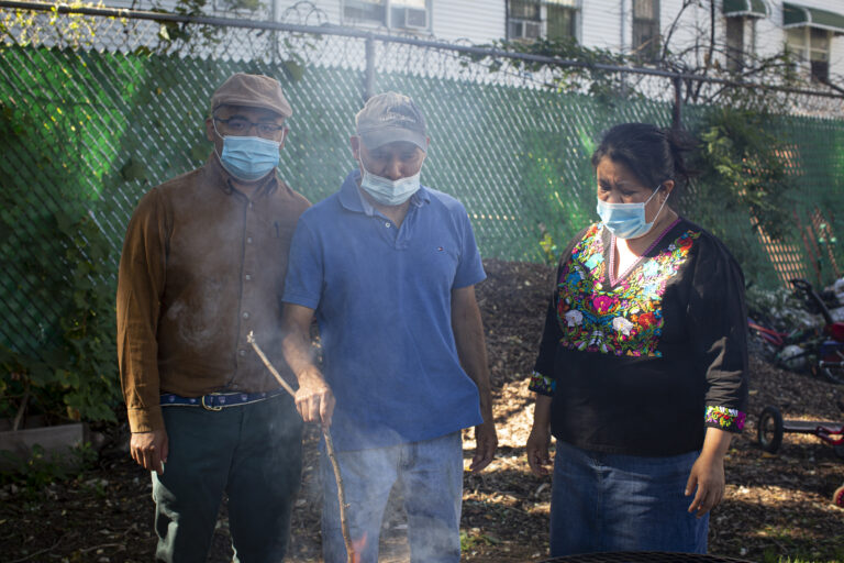 Marco, wearing a light brown shirt, jeans, and a hat, Antonio, wearing a light blue shirt, jeans, and a baseball cap, and Natalia, wearing an Indigenous Mexican embroidered shirt (black with flowers) and a long denim skirt, stand near the fire (out of frame). All three are wearing medical masks. Smoke comes out of the fire. The mounds of soil in the background reflect the morning sunlight. Photo by Cinthya Santos-Briones for Brewing Memories workshop, October 3, 2020.