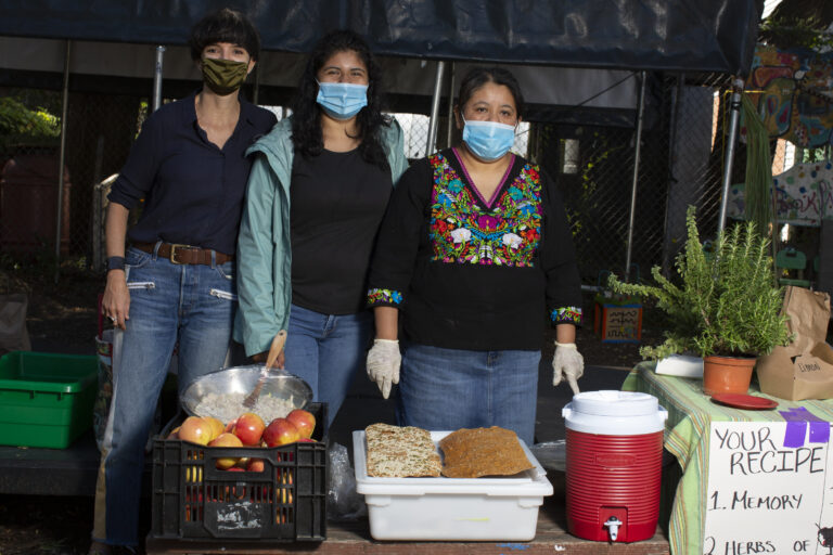Angeles, wearing a dark blue shirt and jeans, Carolina, wearing a light green jacket, a black shirt and jeans, and Natalia, wearing an Indigenous Mexican embroidered shirt (Black with flowers) and a long denim skirt, pose for a photo behind a crate full of red apples, a tray with bread and a big red container with hot water. The three of them are wearing masks. On the right side there is a table with a rosemary plant and a poster with instructions that reads “your recipe” “memory” and “herbs.” Photo by Cinthya Santos-Briones for Brewing Memories workshop, October 3, 2020.