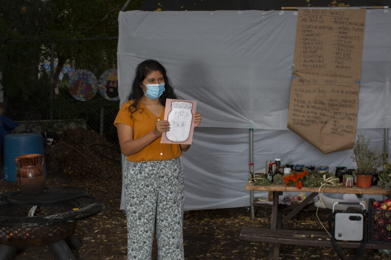 Carolina, wearing an orange shirt and patterned pants, shows a drawing to the participants of the workshop (out of frame). She wears a medical mask. She stands to the right of the table showcasing the different plants participants will learn about during the workshop. On the bench there is a crate with apples and a speaker. In the back, a paper kraft board shows a list of words in English and Spanish that Carolina uses as part of the activity. Photo by Cinthya Santos-Briones for Brewing Memories workshop, October 24, 2020.