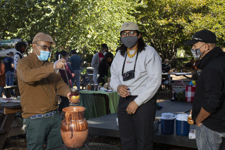 Marco, who is wearing a light brown shirt, green pants, and hat and medical mask, stands near a fire, serving tea to workshop participants of different ages, who stand in line, waiting to be served; the two people in line are also wearing masks––one of whom wears black glasses sunglasses over his gray mask. There’s a big pot made out of clay on top of the fire; smoke comes out of the fire. In the background there are other workshops participants. Photo by Cinthya Santos-Briones for Brewing Memories workshop, October 3, 2020.