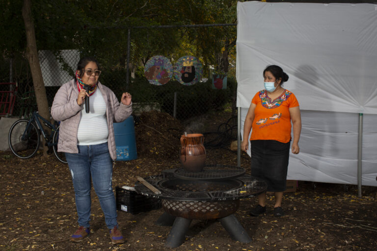 Yajaira, wearing a white shirt, a soft pink jacket, a scarf, and jeans, shares a memory with the participants of the workshop (out of frame). She stands near the fire. Natalia stands near Yajaira, to her left, wearing an Indigenous Mexican embroidered shirt (orange with flowers), a long black skirt, and a medical mask. Photo by Cinthya Santos-Briones for Brewing Memories workshop, October 24, 2020.