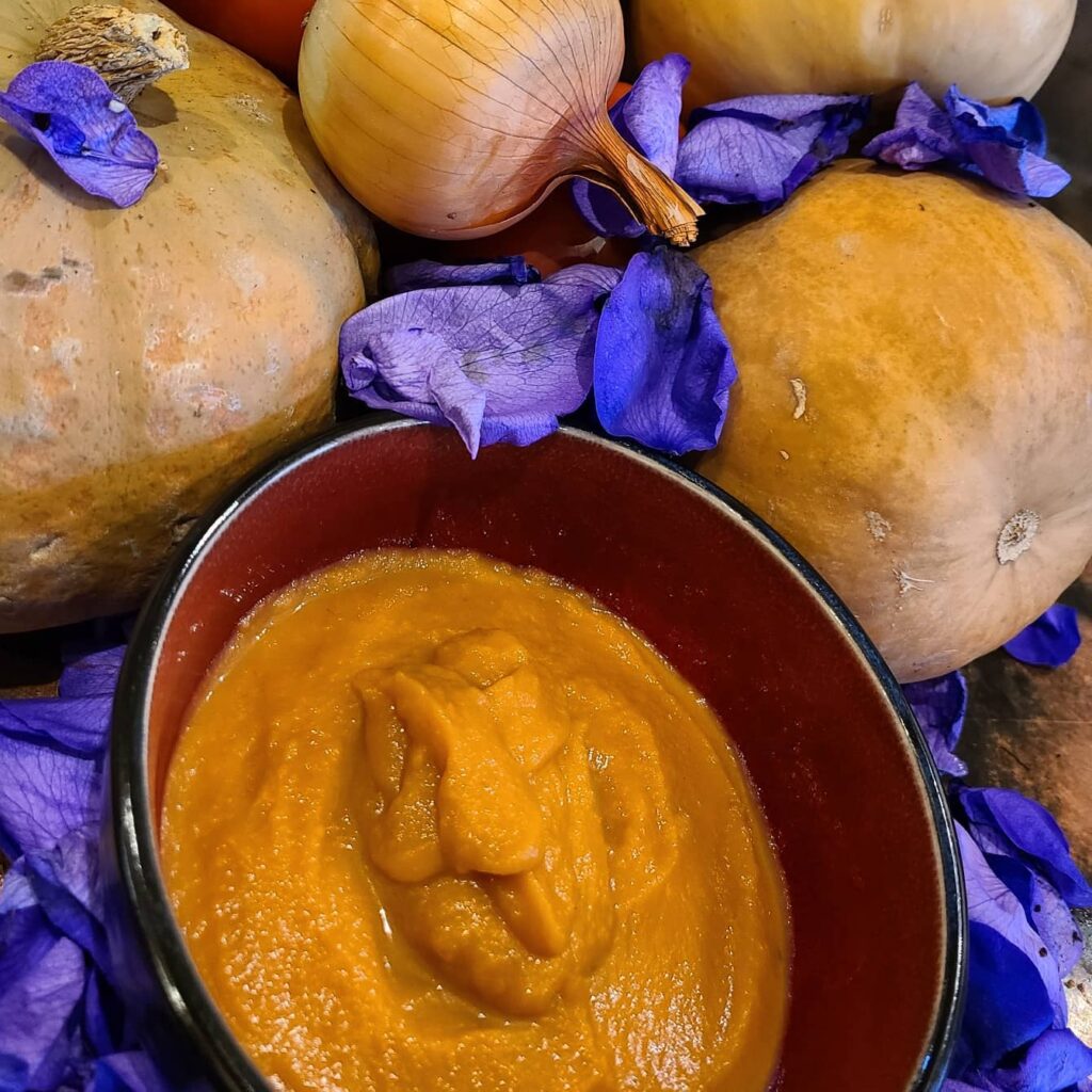 A bowl of pumpkin soup surrounded by whole potatoes and leaves of purple irises. 